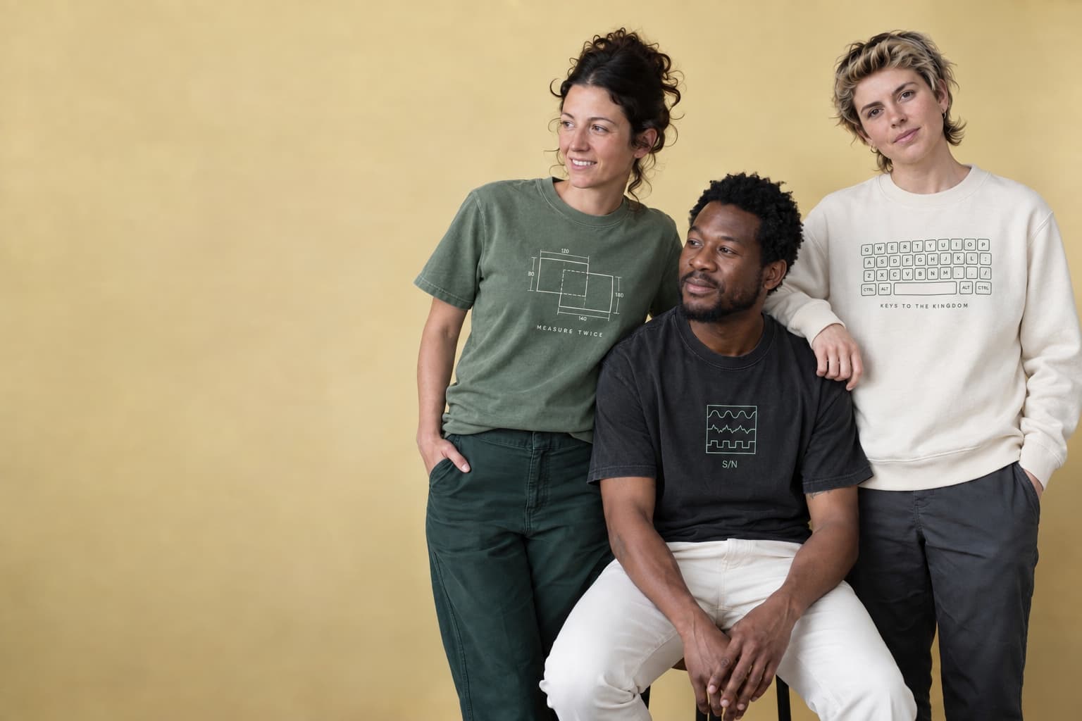 Three creatives wearing the sage Measure Twice tee, charcoal Signal / Noise tee, and cream Keys to the Kingdom crewneck